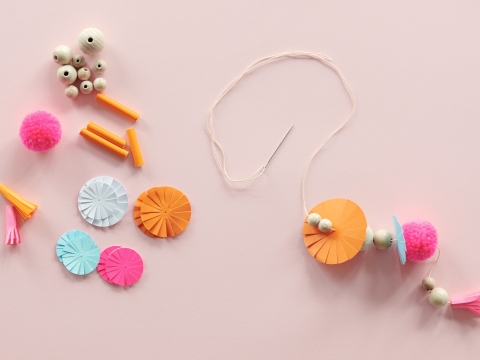 Alle Teile bereitlegen: Holzperlen, Papierblumen, Papierzottel, Pompons. Den Faden durch die Nadel ziehen und nach Lust und Laune auffädeln. Mindestens 8 längere und 4 kürzere Fäden (für oben) herstellen.