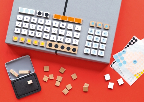 Kleine Kartonstücke zuschneiden, Buchstaben und Zahlen darauf stempeln oder kleben. Die einzelnen «Tasten» auf dem Kartonschachteldeckel platzieren und festkleben.
